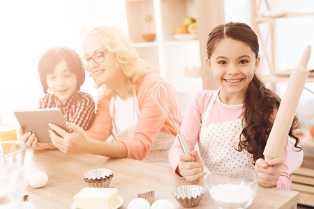 Little girl holds rolling pin and whisk in her hands, sitting in kitchen with her grandmother and...