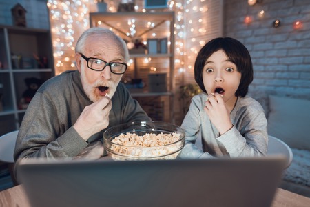 Grandfather and grandson are watching movie with popcorn at table at night at home.の写真素材