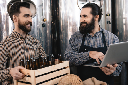 Bearded brewer in apron looks at merry man holding box of empty bottles. Brewing. Brewery. Beer crafting. Process of beer manufacturing.の写真素材