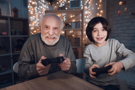 Grandfather and grandson are playing video games at table at night at home.の写真素材