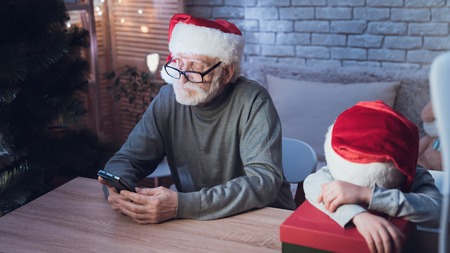 Grandfather and grandson in Santa Claus's hats at table at night at home. Granddad is on phone, boy is sleeping.の写真素材