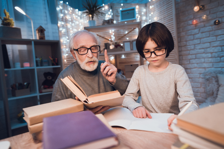 Grandfather and grandson are doing homework at table at night at home. Granddad is helping boy.の写真素材