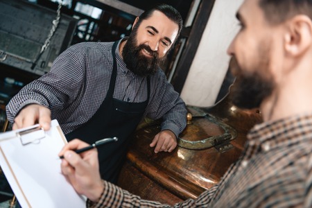 Contented bearded brewer in apron gives documents for signing man in shirt in brewery. Process of beer manufacturing. Brewing. Brewery.の写真素材