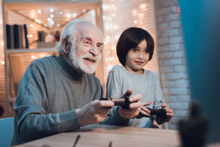 Grandfather and grandson are playing video games on computer at table at night at home.