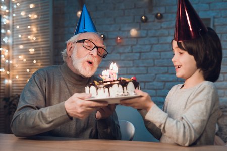Grandfather and grandson at table at night at home. Birthday party. Granddad is giving boy birthday cake.の写真素材