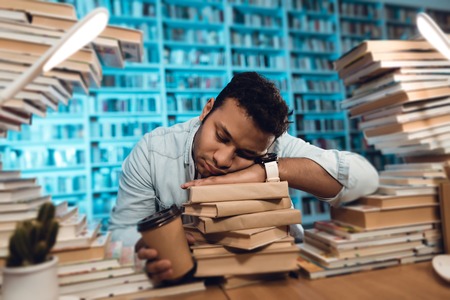 Ethnic indian mixed race guy sitting at table surrounded by books in library. Student is sleeping.の写真素材