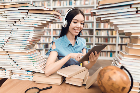 Ethnic asian girl sitting at table surrounded by books in library. Student is using tablet.の写真素材