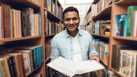 Ethnic indian mixed race student in book aisle of library surrounded by books.