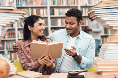Ethnic indian mixed race girl and guy sitting at table surrounded by books in library. Students are reading book.の写真素材