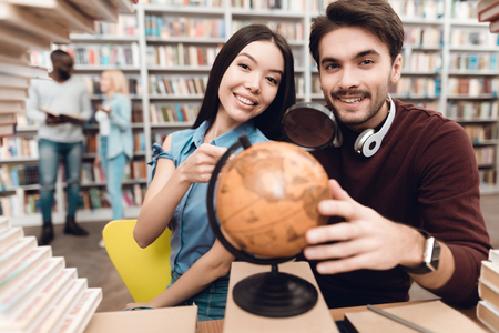 Ethnic asian girl and white guy sitting at table surrounded by books in library. Students are using globe.の写真素材