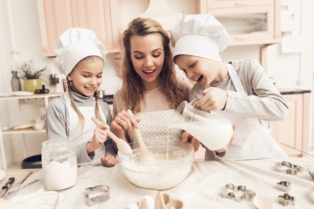 Children in chef's hats with mother in kitchen. Mother is stirring mix and brother is adding milk.