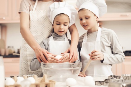 Children in chef's hats with mother in kitchen. Mother is teaching kids how to break eggs.