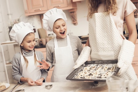 Children in chef's hats with mother in kitchen. Mother is holding tray with cookies.の写真素材
