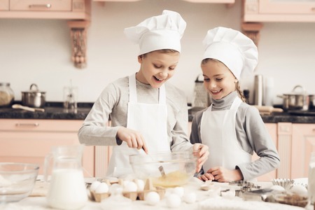 Children in chef's hats in kitchen. Brother and sister are whisking eggs with fork.の写真素材