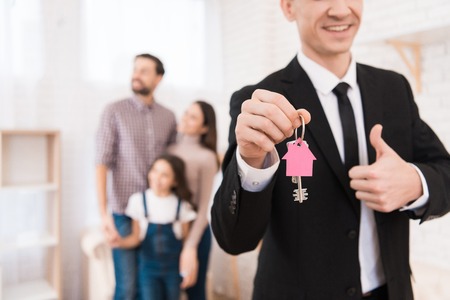 Realtor in suit holds keys with key fob in form of house. Young family looks apartment. Buying real estate concept.の写真素材