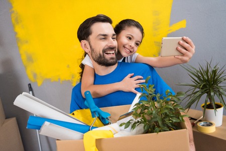 Little daughter using smartphone does selfie with her father who keeps box of tools and things....