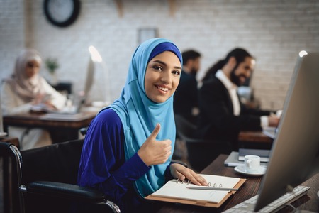 Disabled arab woman in hijab in wheelchair working in office. Woman is giving thumbs up.の写真素材