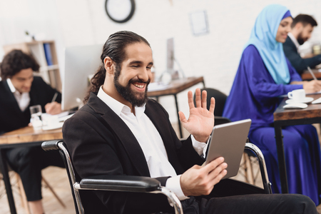 Disabled arab man in suit in wheelchair working in office. Man is talking on tablet.の写真素材