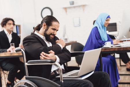 Disabled arab man in suit in wheelchair working in office. Man's elbow hurts.の写真素材
