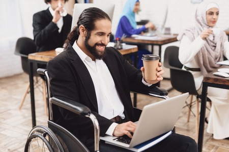 Disabled arab man in suit in wheelchair working in office. Man is working on laptop and drinking...
