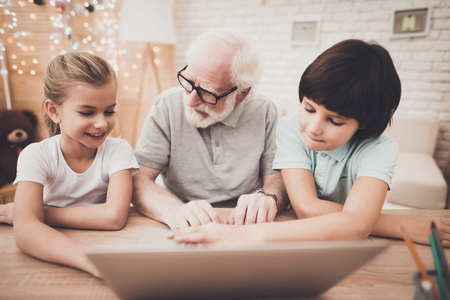 Grandfather, grandson and granddaughter at table at home. Grandpa and children are using laptop.