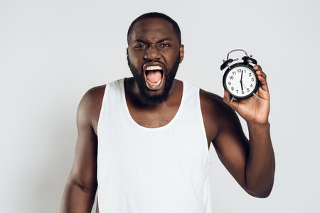 Angry African American man yells holding alarm clock. Morning awakening. Waking up. Isolated on white background. Studio portrait.の写真素材