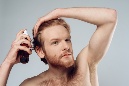 Red haired hairy man sprinkles balm on hair. Hair care. Isolated on grey background. Studio portrait. Male beauty concept.の写真素材