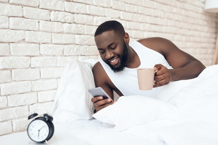 Black, awakened man drinks coffee in bed browsing web. Early morning. Pleasant awakening.の写真素材