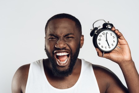 Angry African American man yells holding alarm clock. Morning awakening. Waking up. Isolated on white background. Studio portrait.の写真素材
