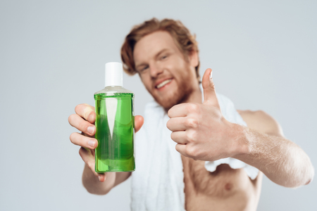 Red haired man posing with bottle of mouthwash and showing thumbs up. Care of teeth. Dental hygiene. Isolated on grey background. Studio portrait.の写真素材