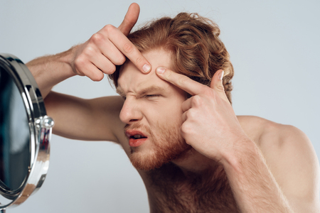 Red haired young man squeeze out pimple on forehead, looking into mirror. Morning hygiene procedures. Isolated on grey background. Studio portrait. Acne. Skin care.の写真素材