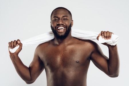Smiling black man with naked torso holds towel. Morning hygiene procedures. Isolated on white background. Studio portrait.の写真素材