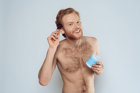 Young happy man is cleaning ears with cotton swab. Morning hygiene procedures. Isolated on grey background. Studio portrait.の写真素材