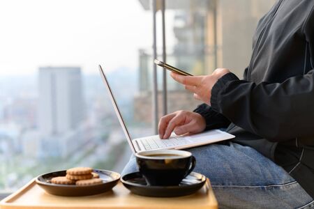 casual wearing man operating smartphone and laptop with coffee and cookies nearby on the balcony...