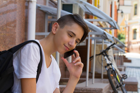 Outside portrait of teen boy. Handsome teenager carrying backpack on one shoulder and smiling, speaking by phoneの写真素材
