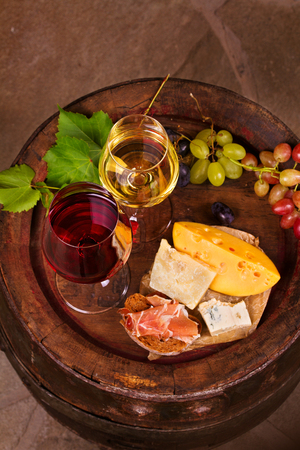 Red and white wine with cheese, prosciutto and grape on old wine barrel in cellar. Food and drinks concept. View from above, top, verticalの写真素材