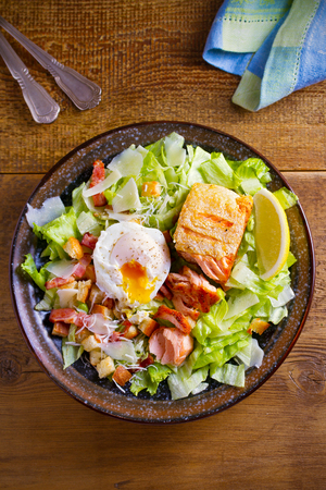 Salmon caesar salad. Crispy pan fried salmon fillet, bacon, poached egg, romaine lettuce and croutons in bowl on wooden table. overhead, verticalの写真素材
