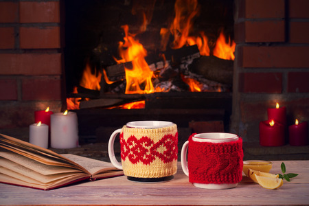 Hot drinks in mugs, book and candles on wooden table beside cosy open fire place. Autumn or winter holidays conceptの写真素材