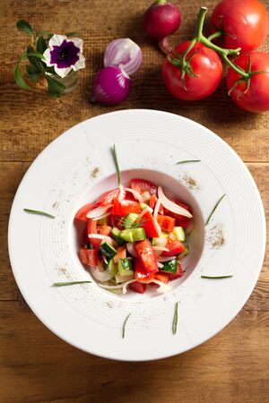 Vegetable salad of fresh tomatoes, cucumbers, bell pepper, onions and herbs in white bowl, wooden table. Overhead, vertical imageの写真素材