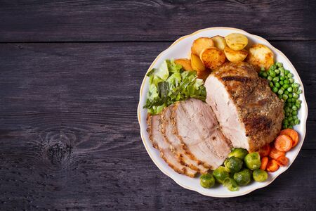 Roasted sliced ham and vegetables: potatoes, carrots, brussel sprouts, cabbage and green peas on black wooden background. copy spaceの写真素材