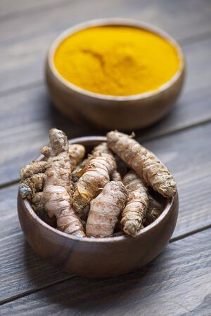 A bowl with turmeric root in the foreground next to bowl with turmeric powder on brown wooden table. Spice for health and ingredient of Indian cuisine.の写真素材
