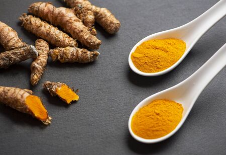 Two spoons with turmeric powder next to turmeric root on black slate base. Spice for health and ingredient of Indian cuisine.の写真素材