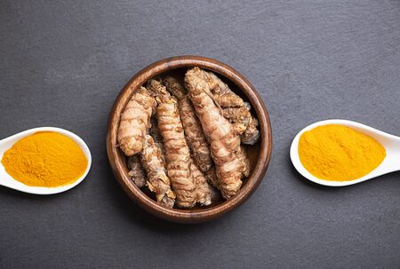 Two white spoons with turmeric powder next to bowl with turmeric root on black slate base. Spice for health and ingredient of Indian cuisine.の写真素材