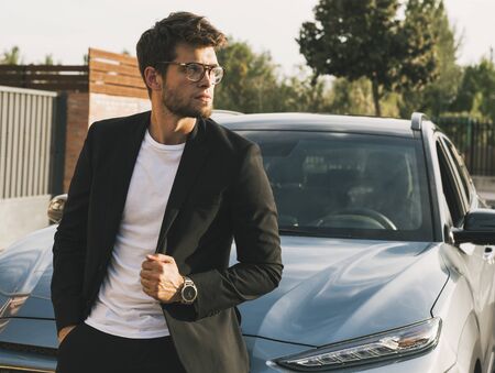 Close-up of attractive young man with beard and glasses in suit is leaning on his car.の写真素材