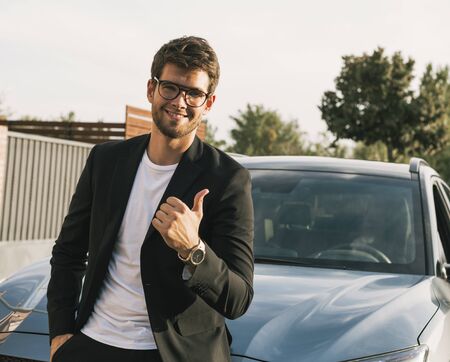 Close-up of attractive young man with beard and glasses in suit is leaning on his car looks at the camera.suitの写真素材