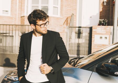 Close-up of attractive young man with beard and glasses in suit is leaning on his car.の写真素材