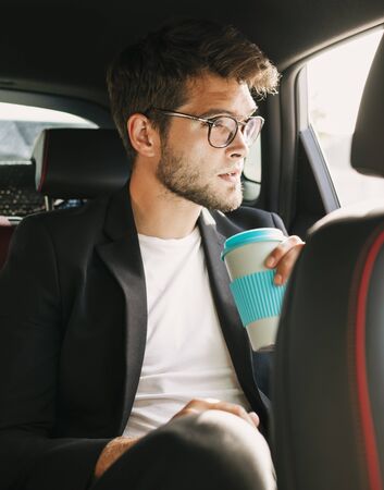 Young and attractive man with a beard and glasses drinks coffee inside a car. Businessの写真素材