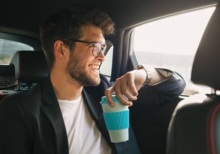 Young and attractive man with a beard and glasses laughs while drinking coffee inside a car. Businessの写真素材
