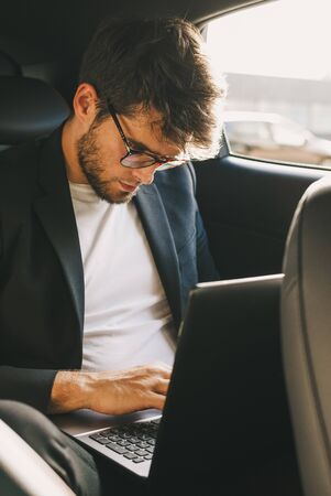 Attractive young man with a beard and glasses works with his laptop inside a car. Businessman.の写真素材