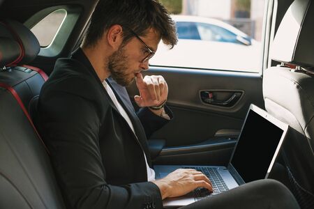 Attractive young man with a beard and glasses works with his laptop inside a car. Businessman.の写真素材